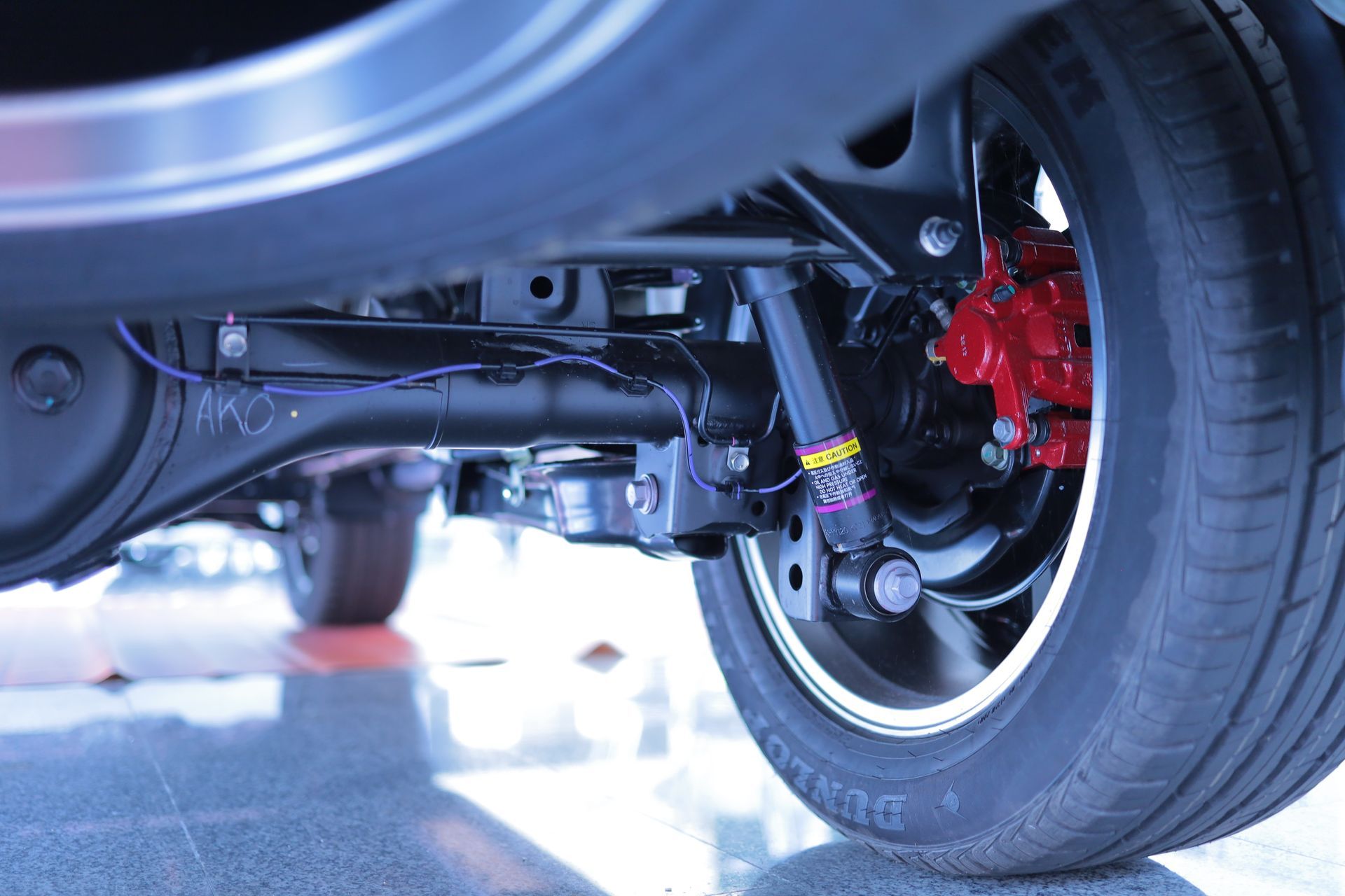 Close-up of a car’s undercarriage and rear wheel with red brake caliper on a garage floor.