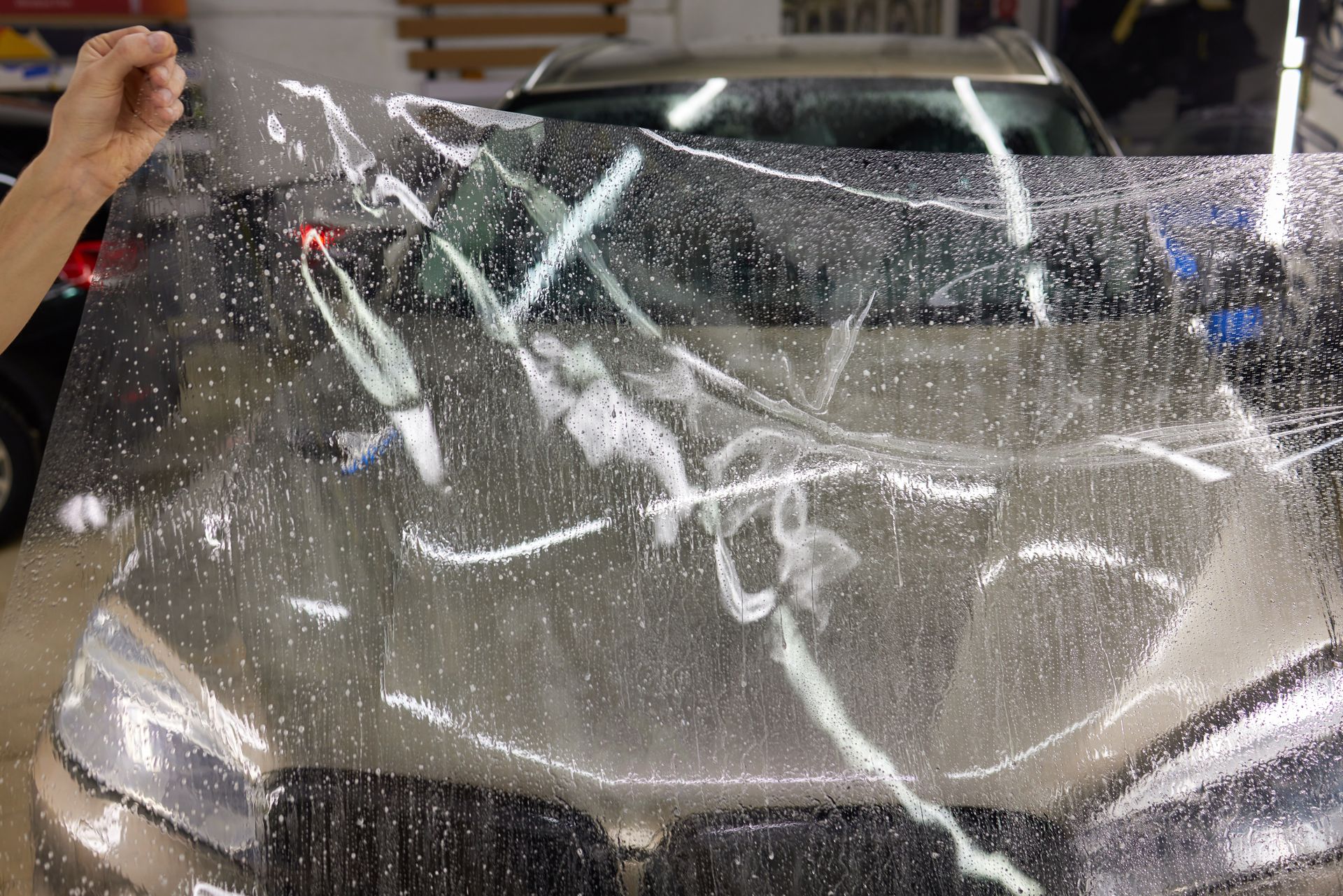 Car covered in a clear protective film, with water droplets and a person peeling it back