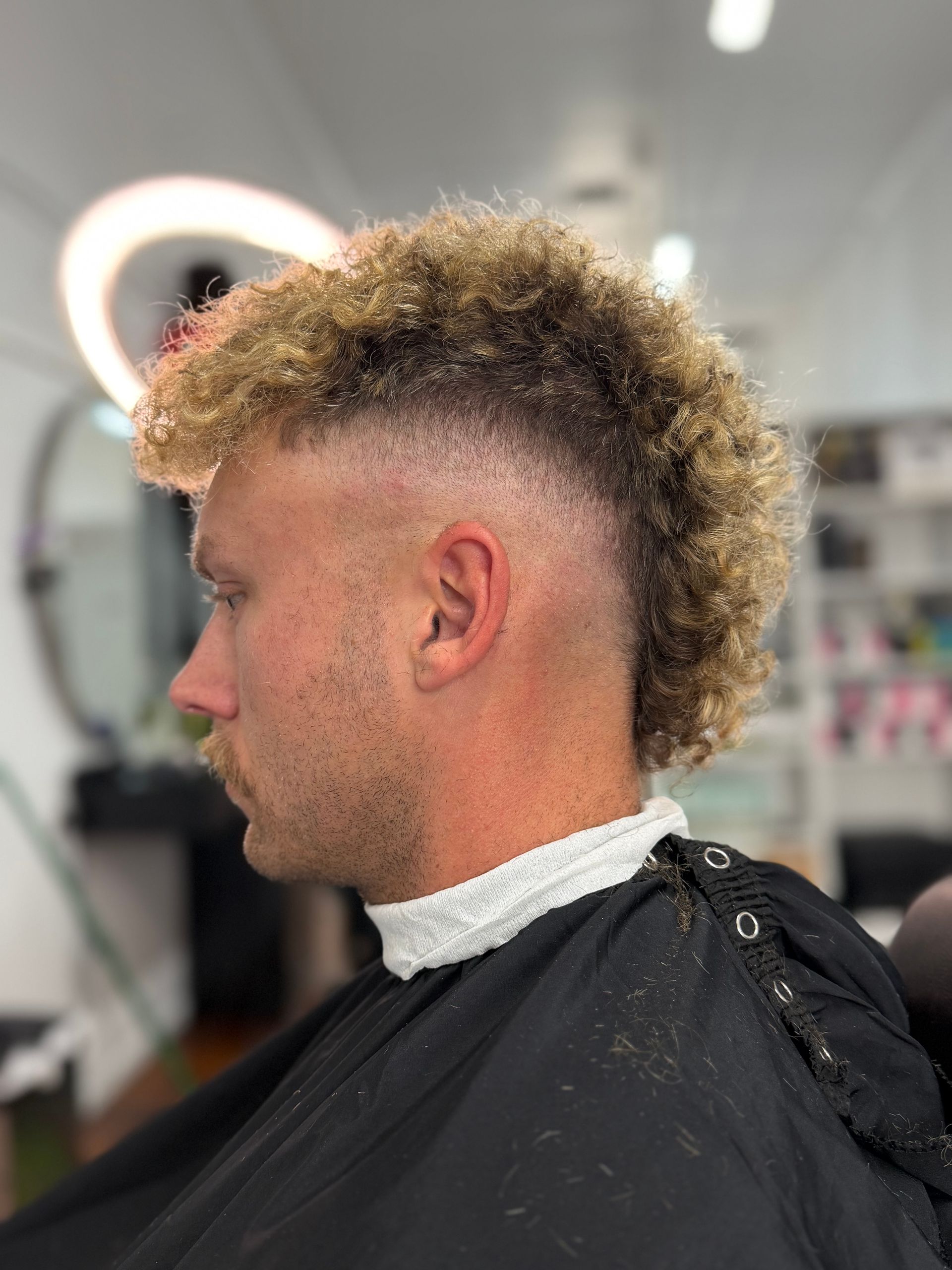 Side profile of a person with a curly mohawk in a barber chair, wearing a cape during a haircut