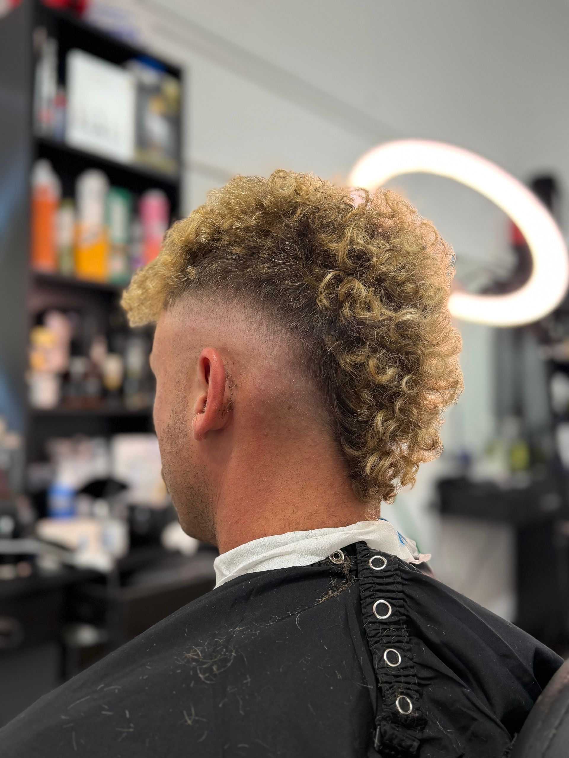 Side-profile haircut with a curly mohawk fade in a barbershop, black cape and white neck strip.