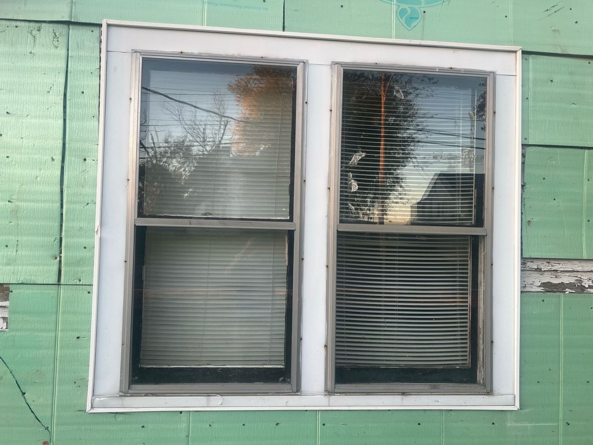 White-framed four-pane window in a green exterior wall with closed blinds.