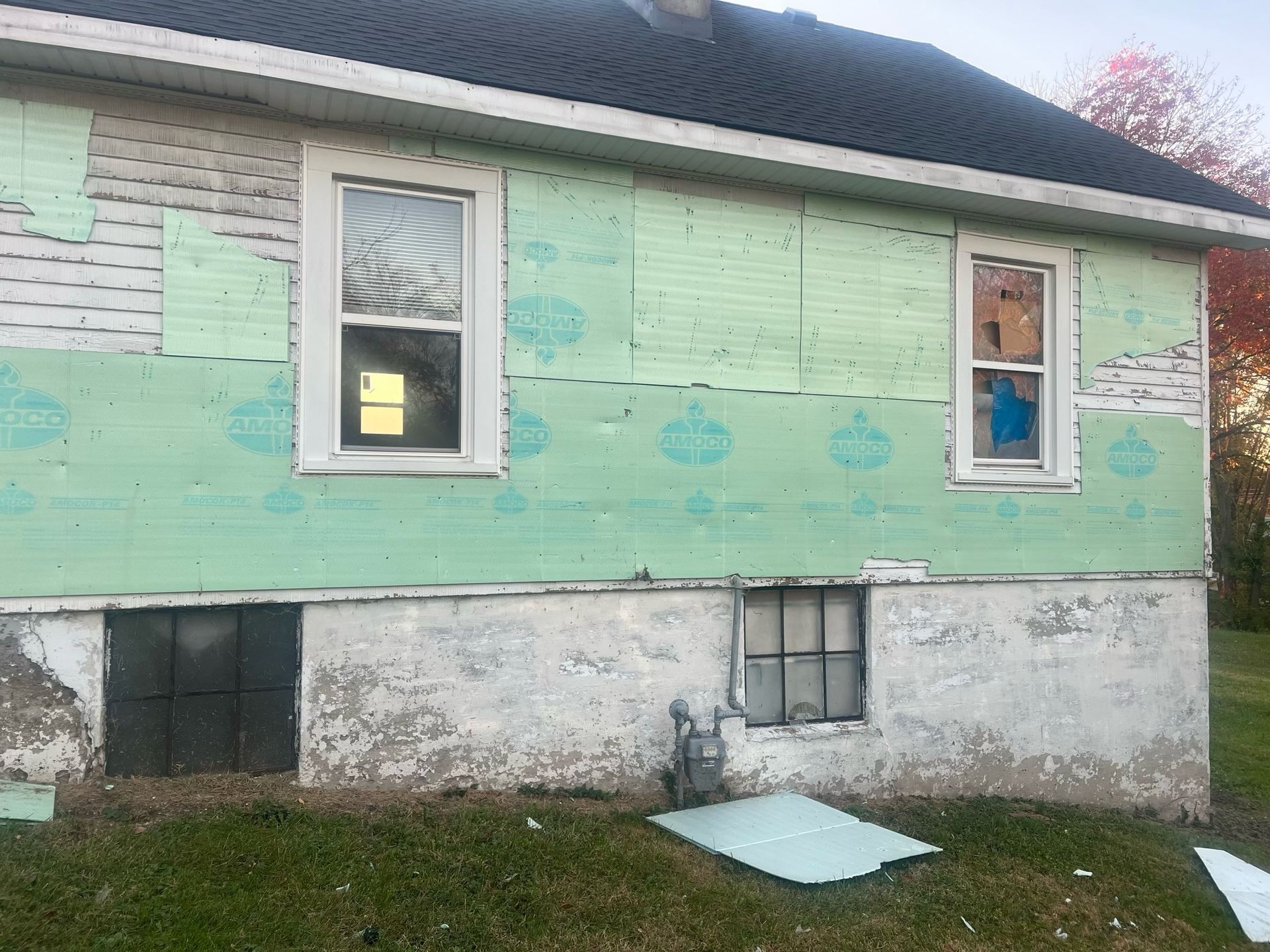 Peeling mint-green house exterior with white lower wall and two windows, one boarded opening near the foundation.