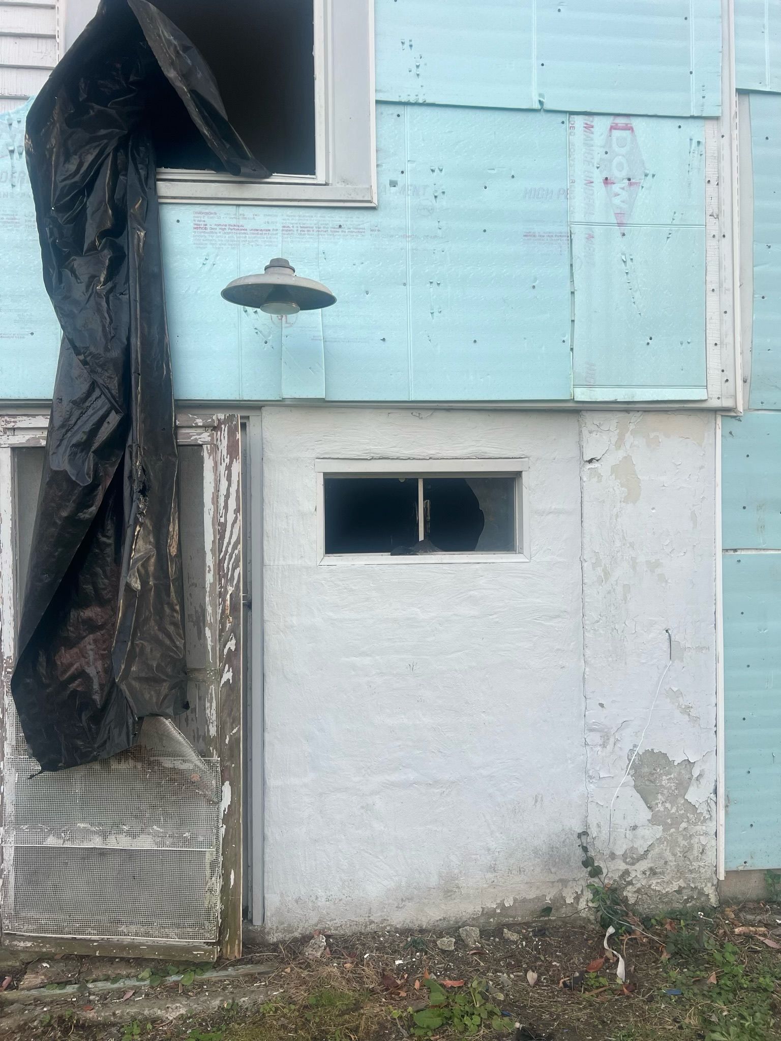 Damaged house exterior with black tarp covering an upper window and broken lower wall opening