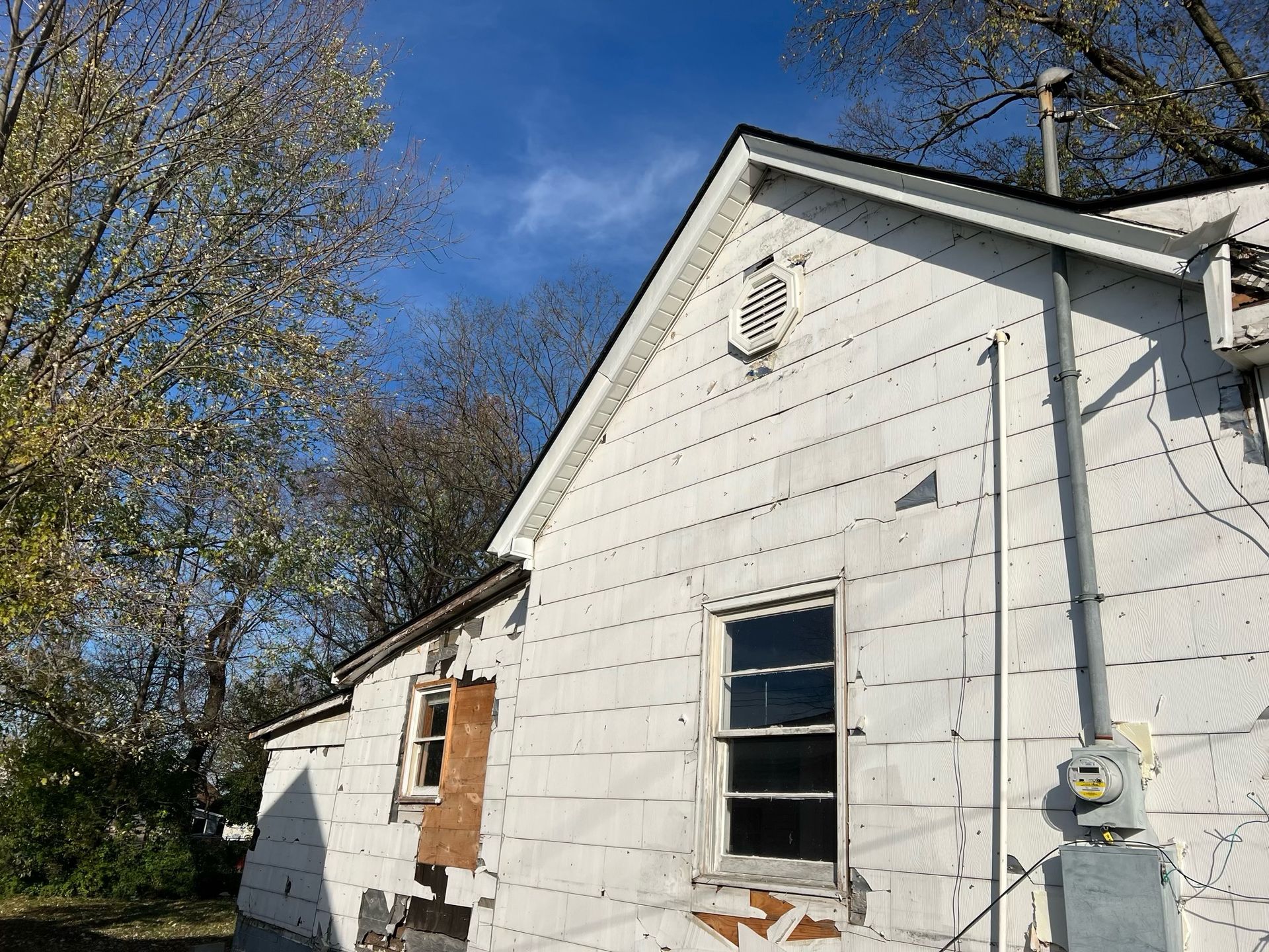 White clapboard house exterior with boarded window, blue sky, and trees in the background