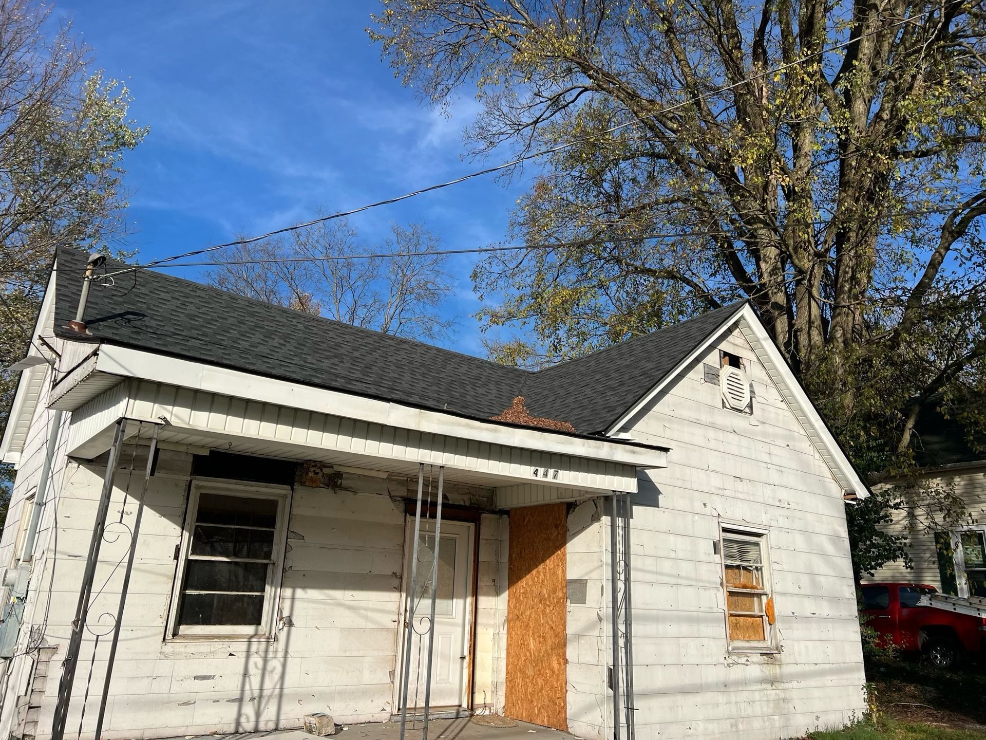 Small white house under repair with boarded windows and a boarded door, trees and blue sky behind it