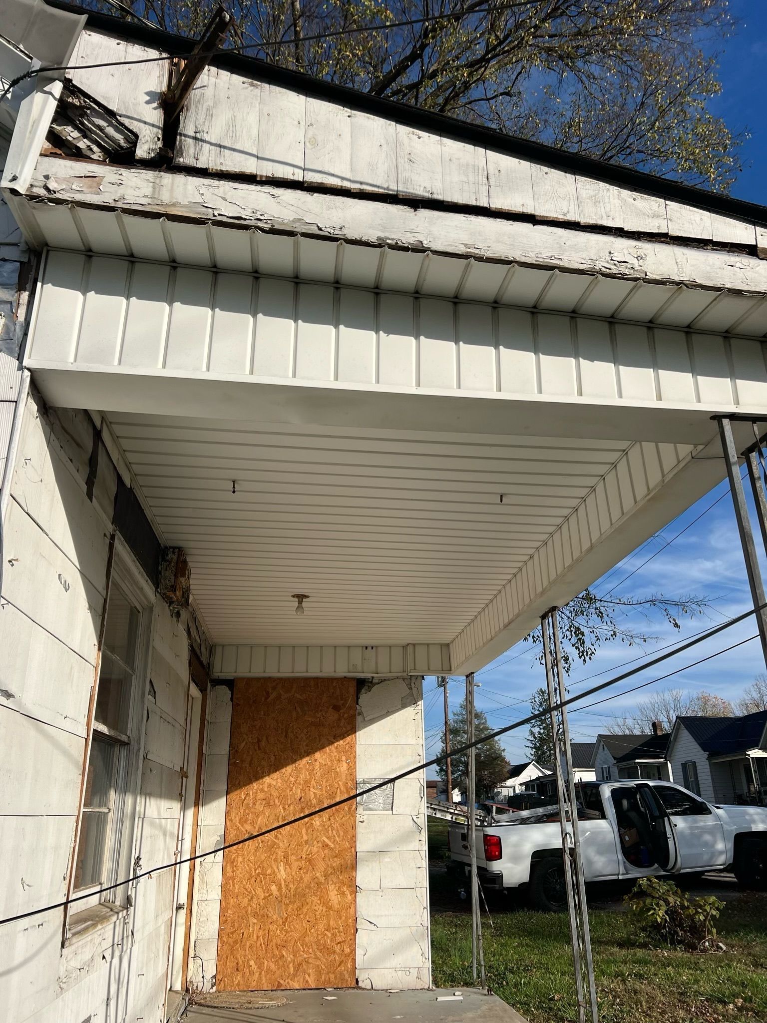 Boarded-up white building entrance with damaged roof and sagging porch, beside a street and utility poles.