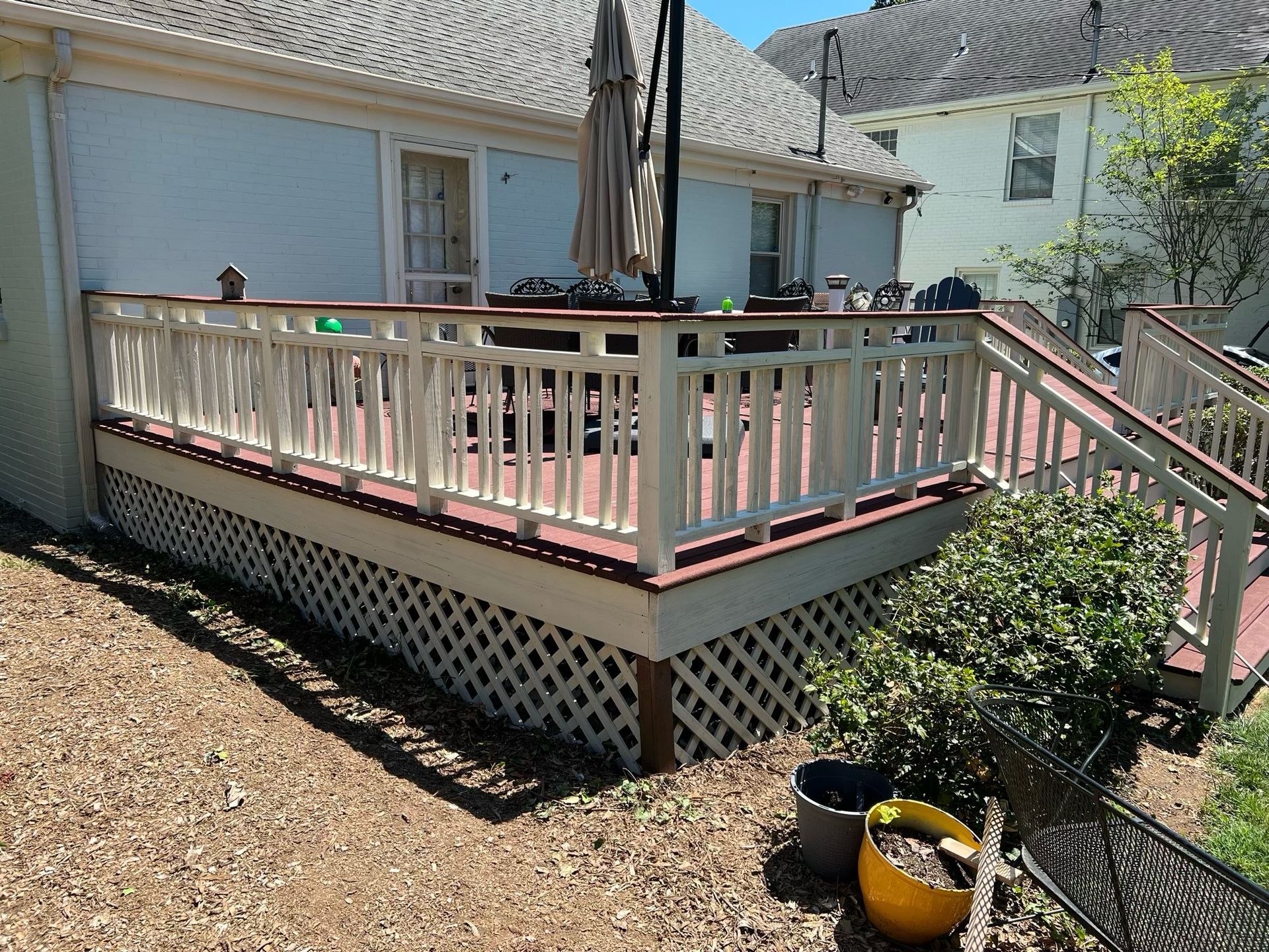 Back deck with pink-and-white railing, steps, closed umbrella, and potted plants beside a house.