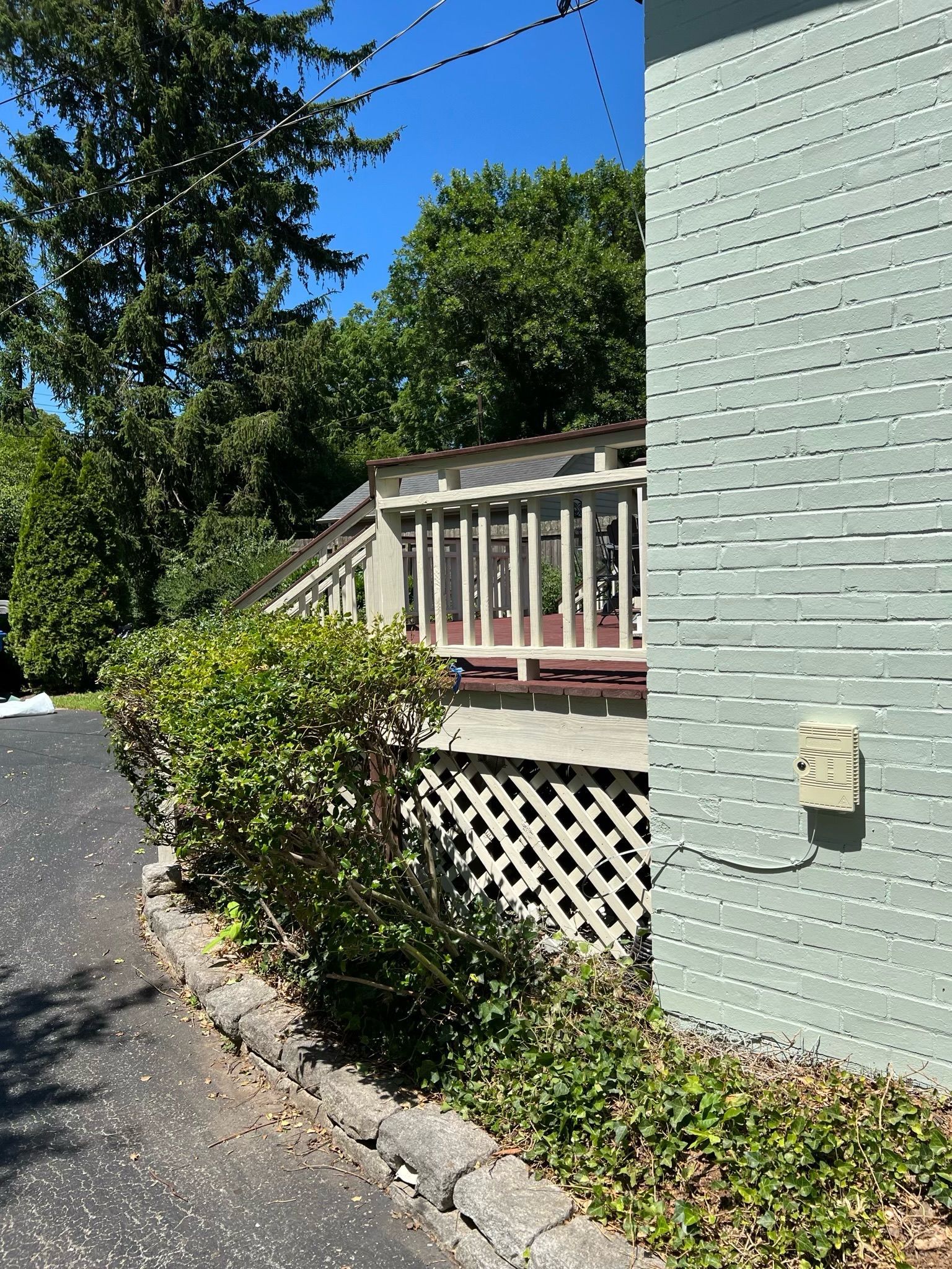 Backyard deck with white railing beside a pale green house, shrubs, and a paved driveway under blue sky
