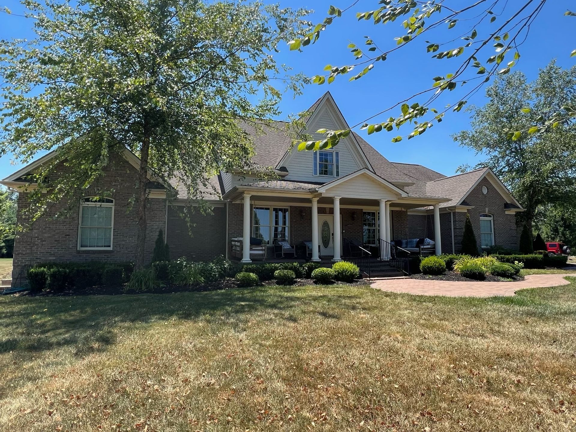 Brick house with a front porch, green lawn, and trees in the yard on a sunny day