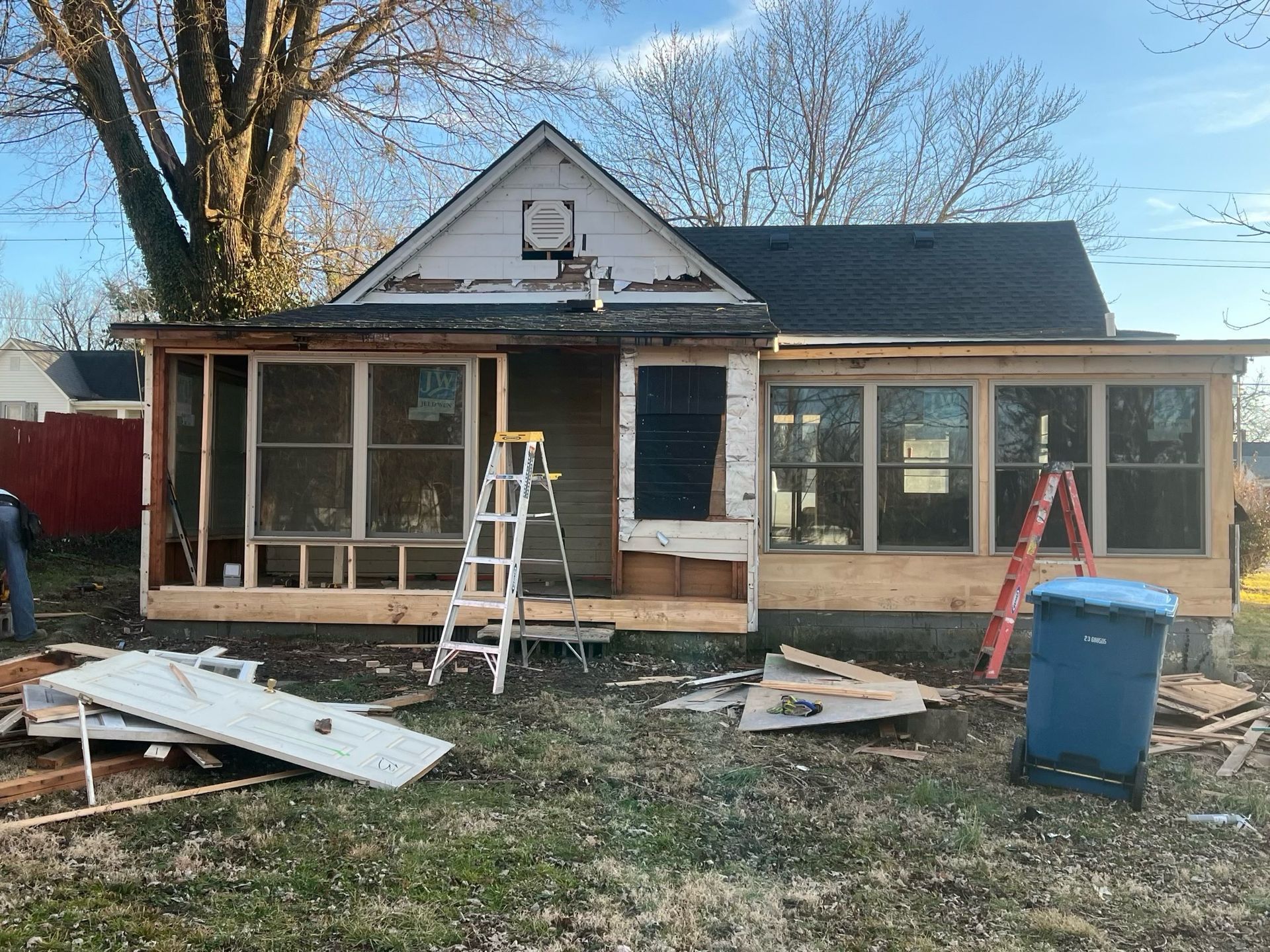 House under renovation with ladders, construction debris, and a blue trash bin in the yard