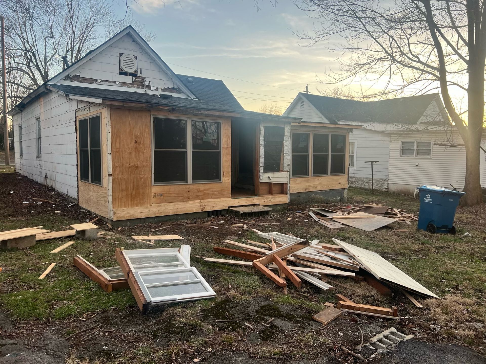 Partially renovated house with plywood-covered windows and construction debris in the yard