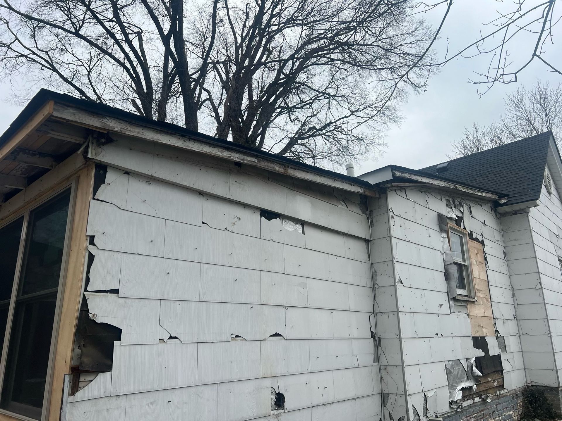 Damaged white house exterior with broken siding and a boarded window under bare tree branches