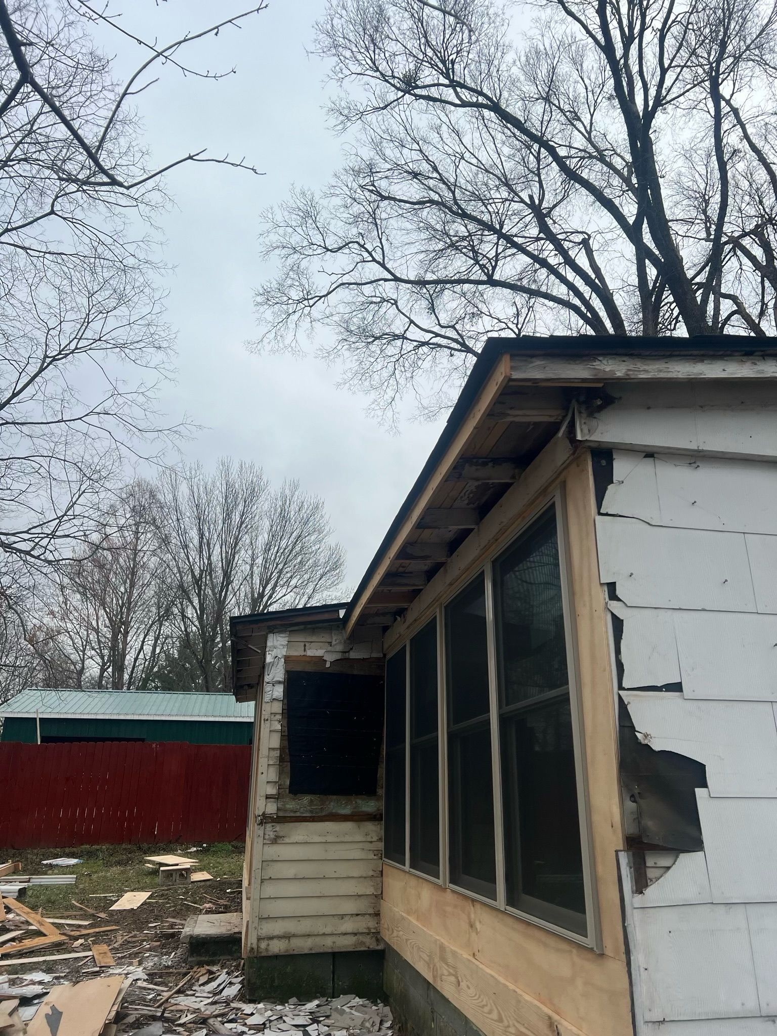 Weathered house exterior with screened porch, broken siding, and leafless trees under an overcast sky