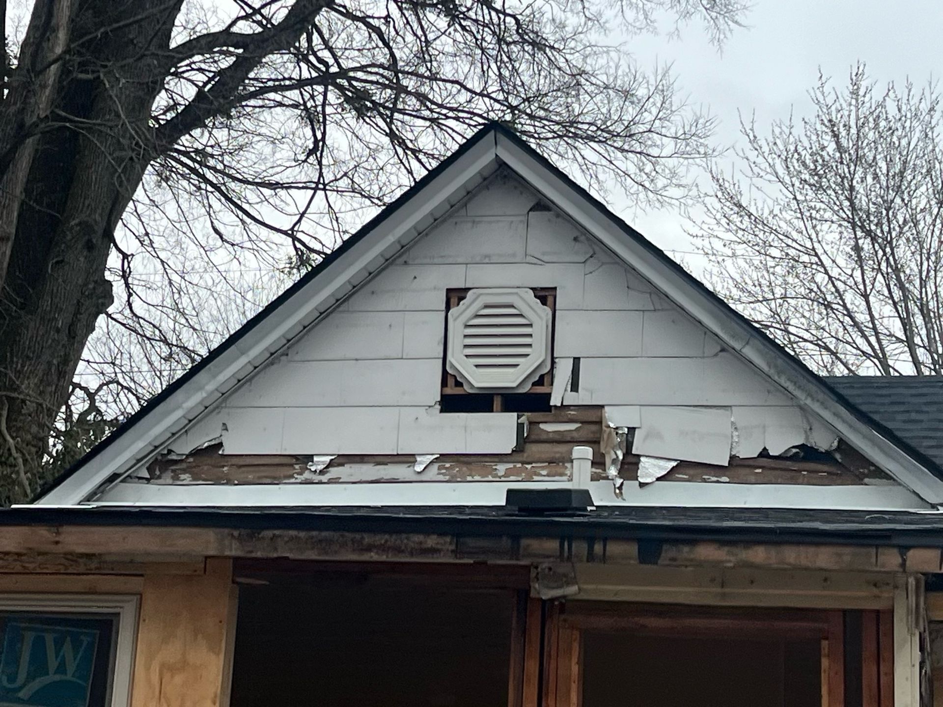 Damaged house roof with missing shingles and a vent under leafless trees