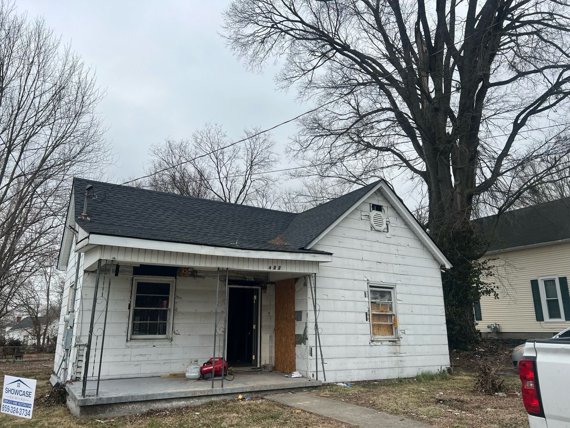 Small white house with boarded windows and an open door, under renovation on a cloudy day.