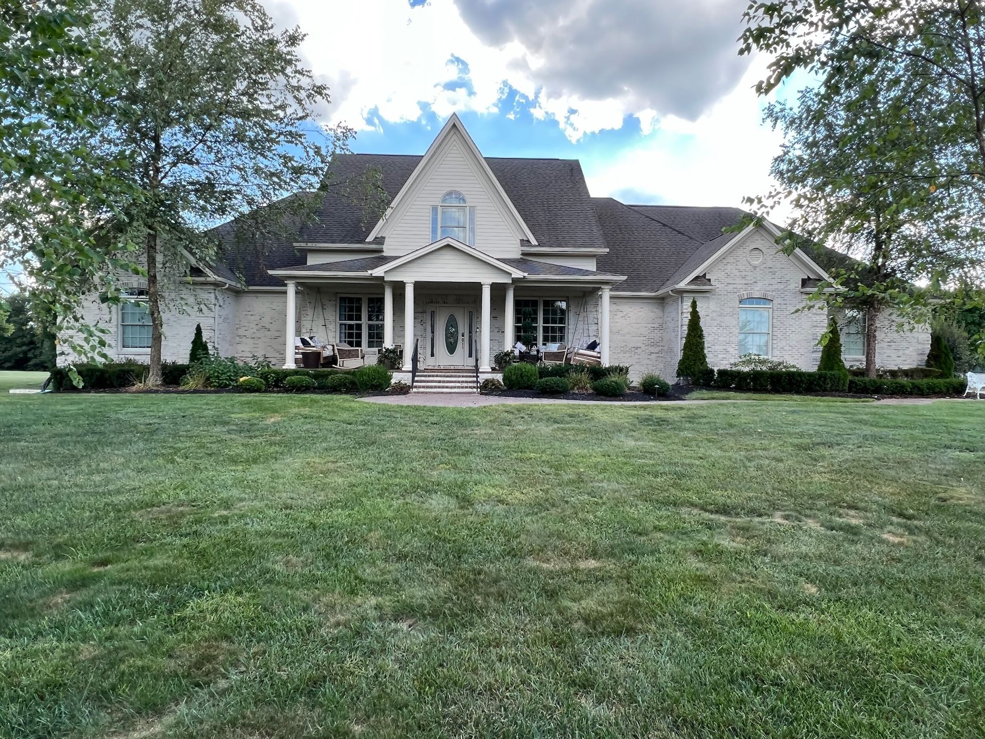 Large light-colored suburban house with a front porch, steep roof, and wide green lawn under a cloudy sky
