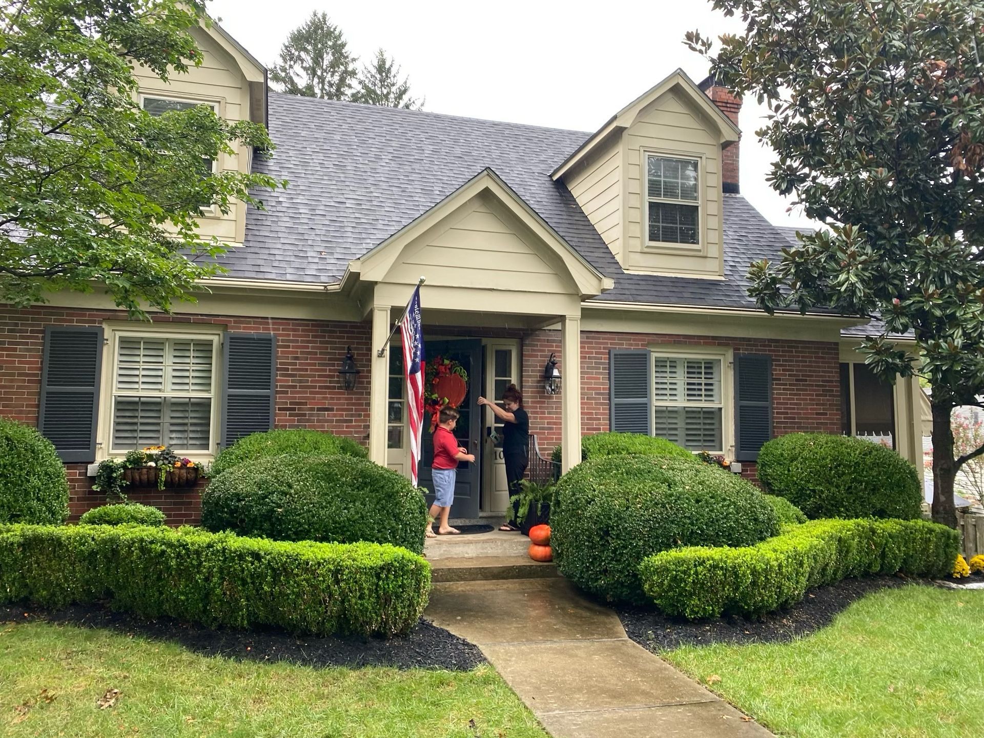 Single-story house with brick facade, trimmed shrubs, and two people standing at the front door.