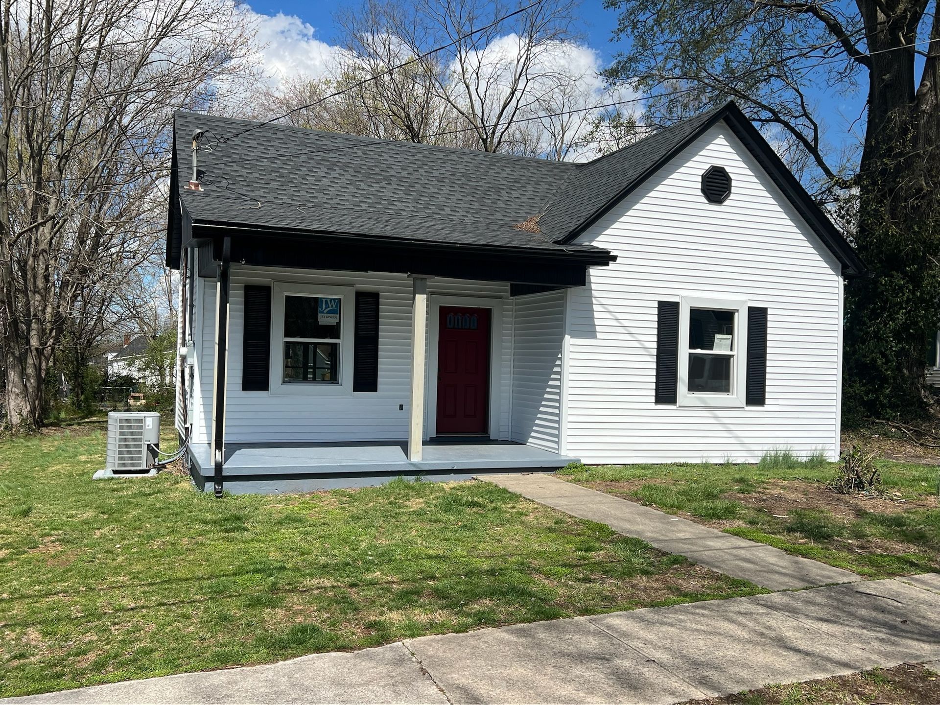 Small white house with black shutters, red door, and a front porch beside a grassy yard.