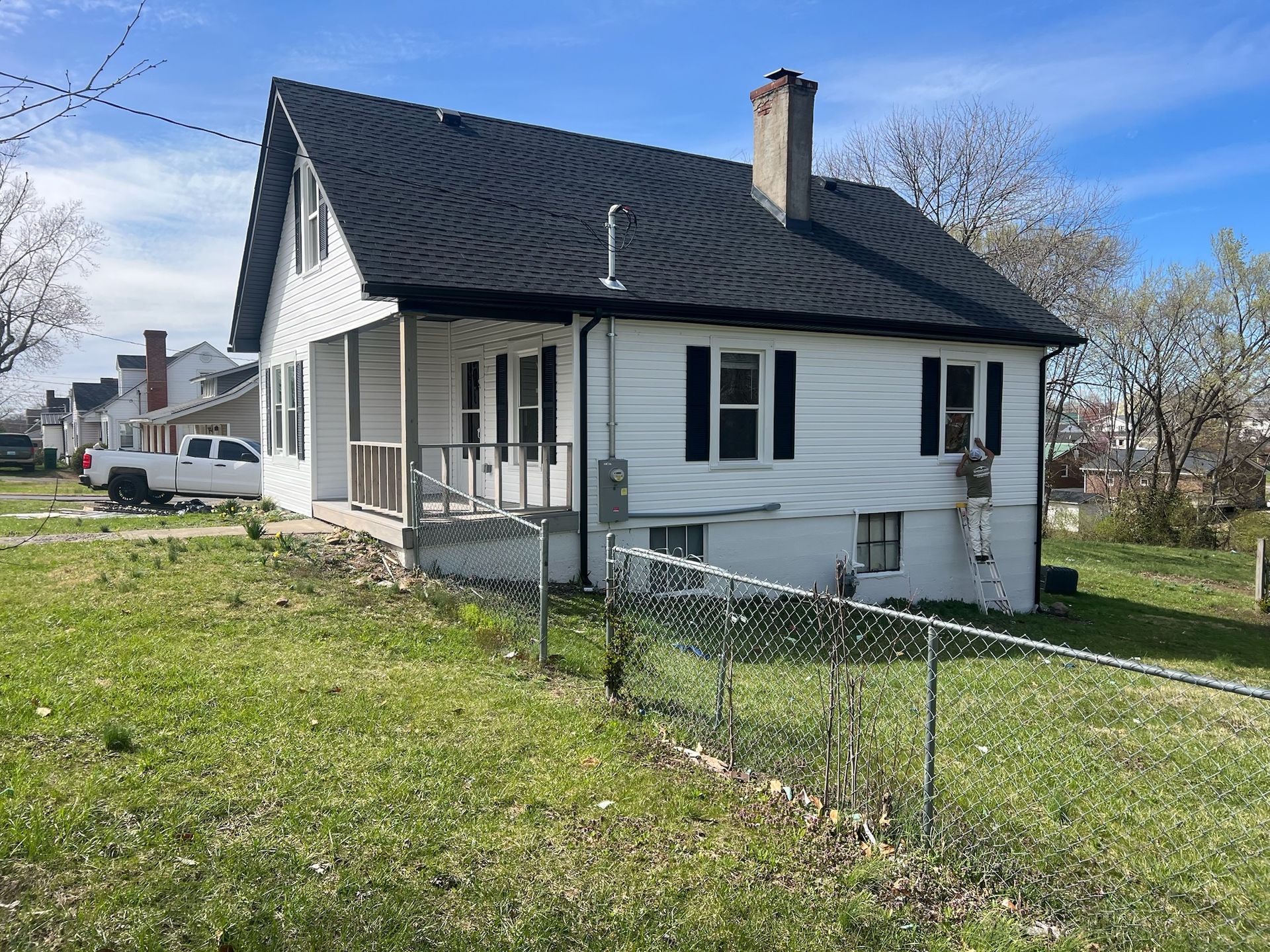 White house with dark roof on a grassy corner lot, with a small porch and chain-link fence