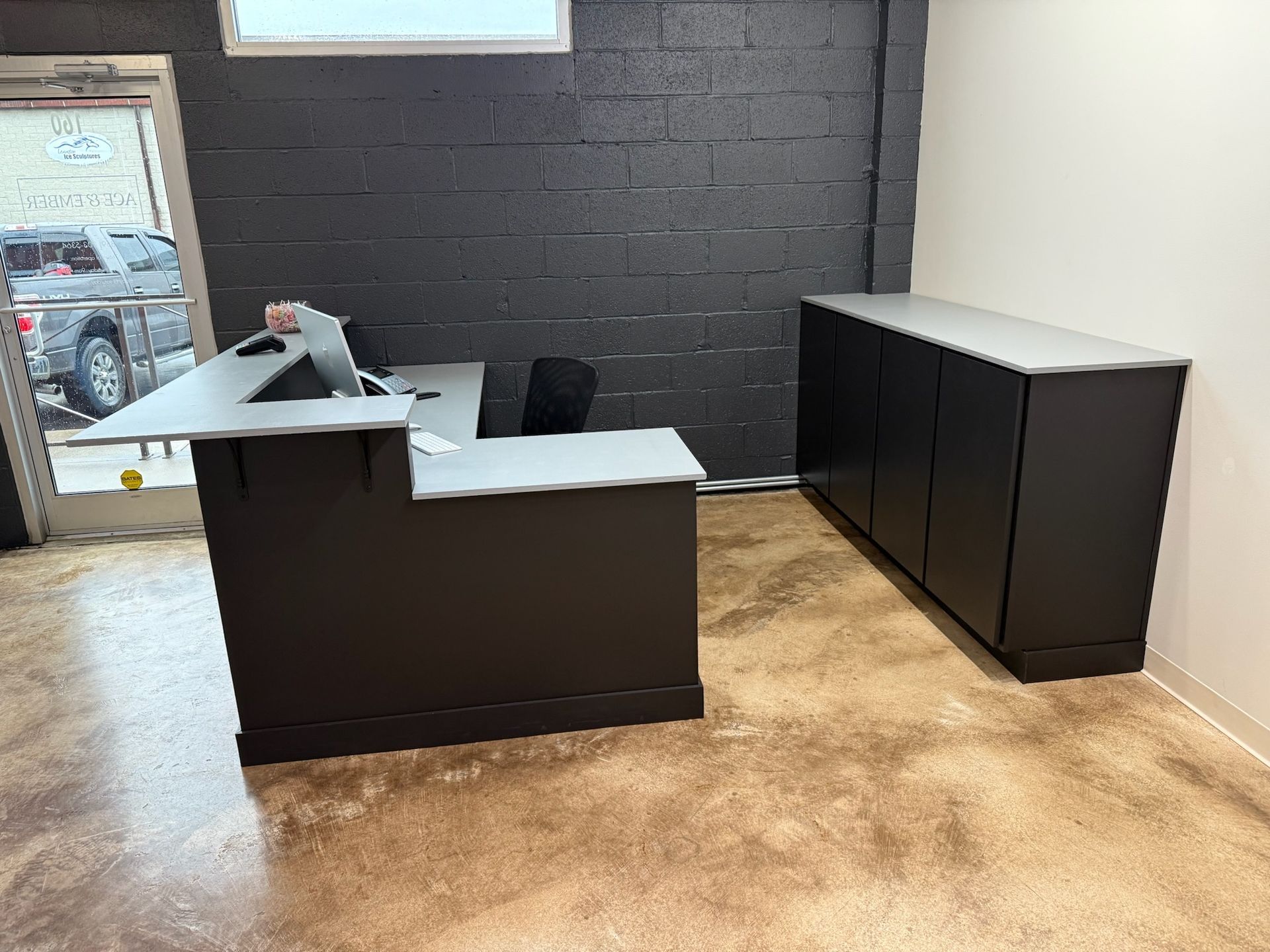 Empty office with a dark desk and cabinets against a black brick wall, tan carpet, and window light.