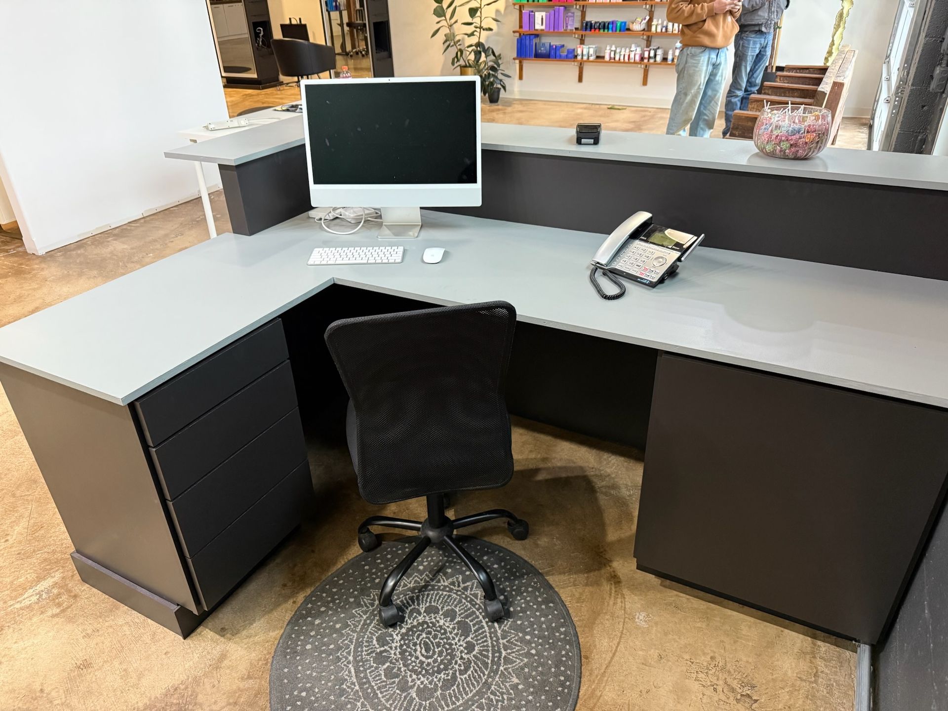 L-shaped gray office desk with black chair, monitor, and phone in a bright workspace