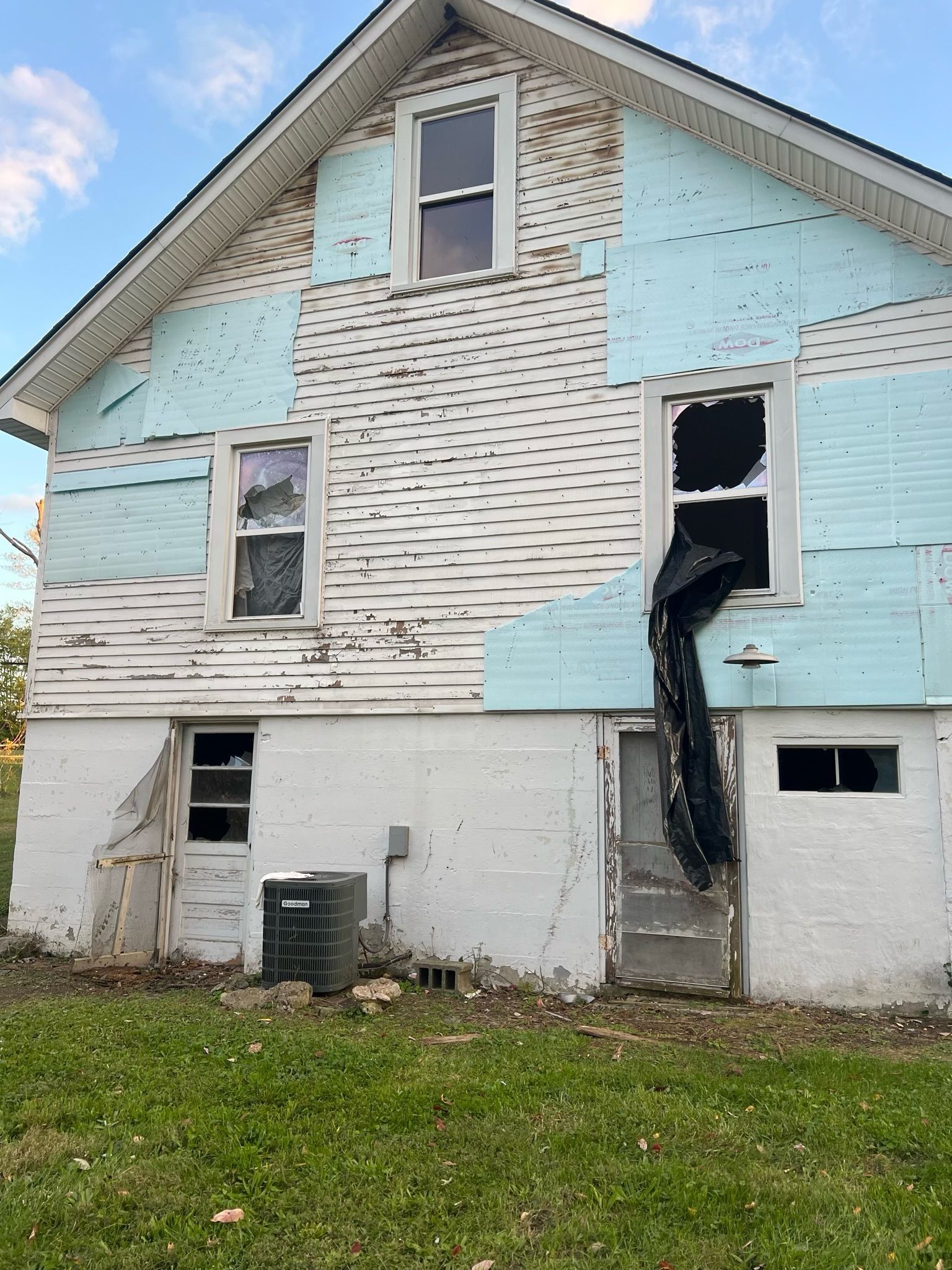 Damaged white and light blue house exterior with broken siding and a black tarp hanging from an upper window