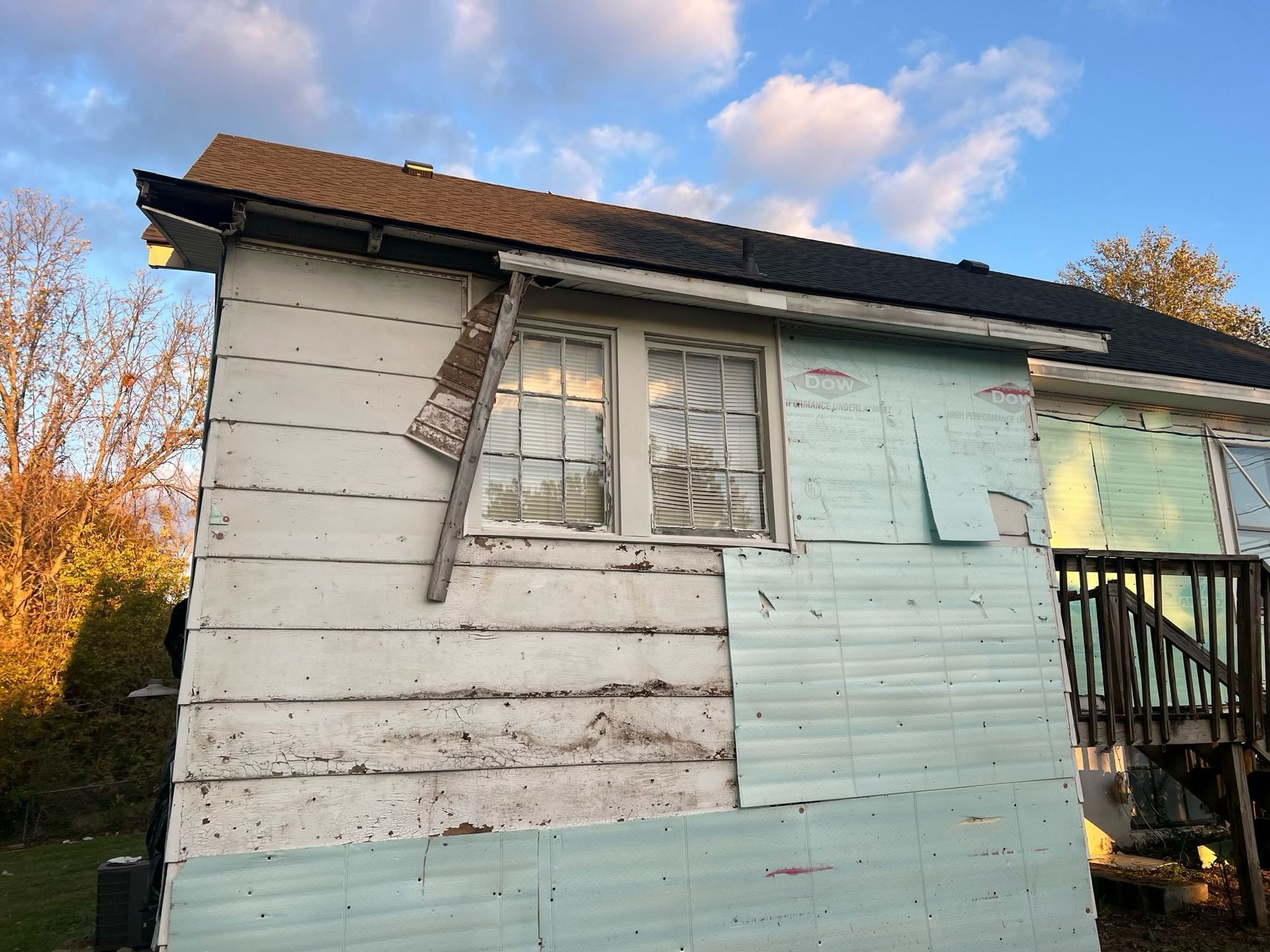 Weathered white house exterior with peeling paint, broken window shutters, and a small porch under a cloudy sky