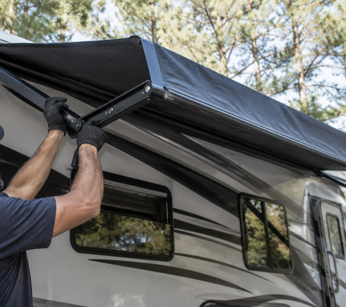 Person opening a black RV awning beside a white camper in a wooded campsite