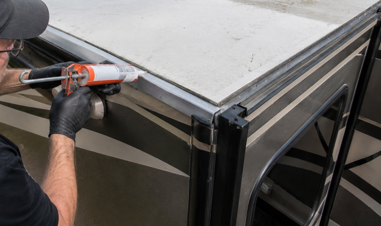 A gloved worker applies sealant with a caulking gun along a black window frame.