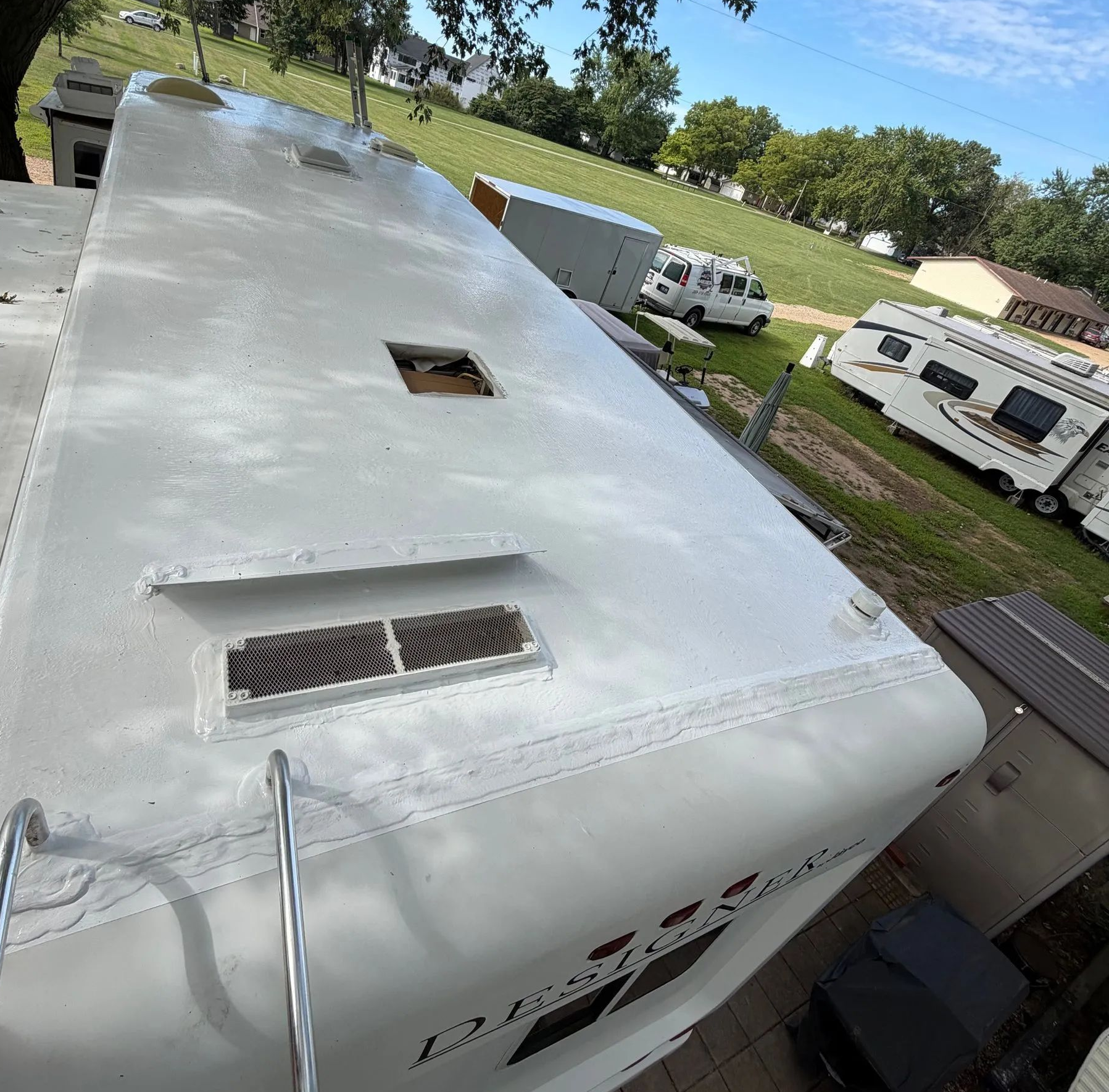 White RV roof with vents and a hatch, viewed from above in a sunny campground.