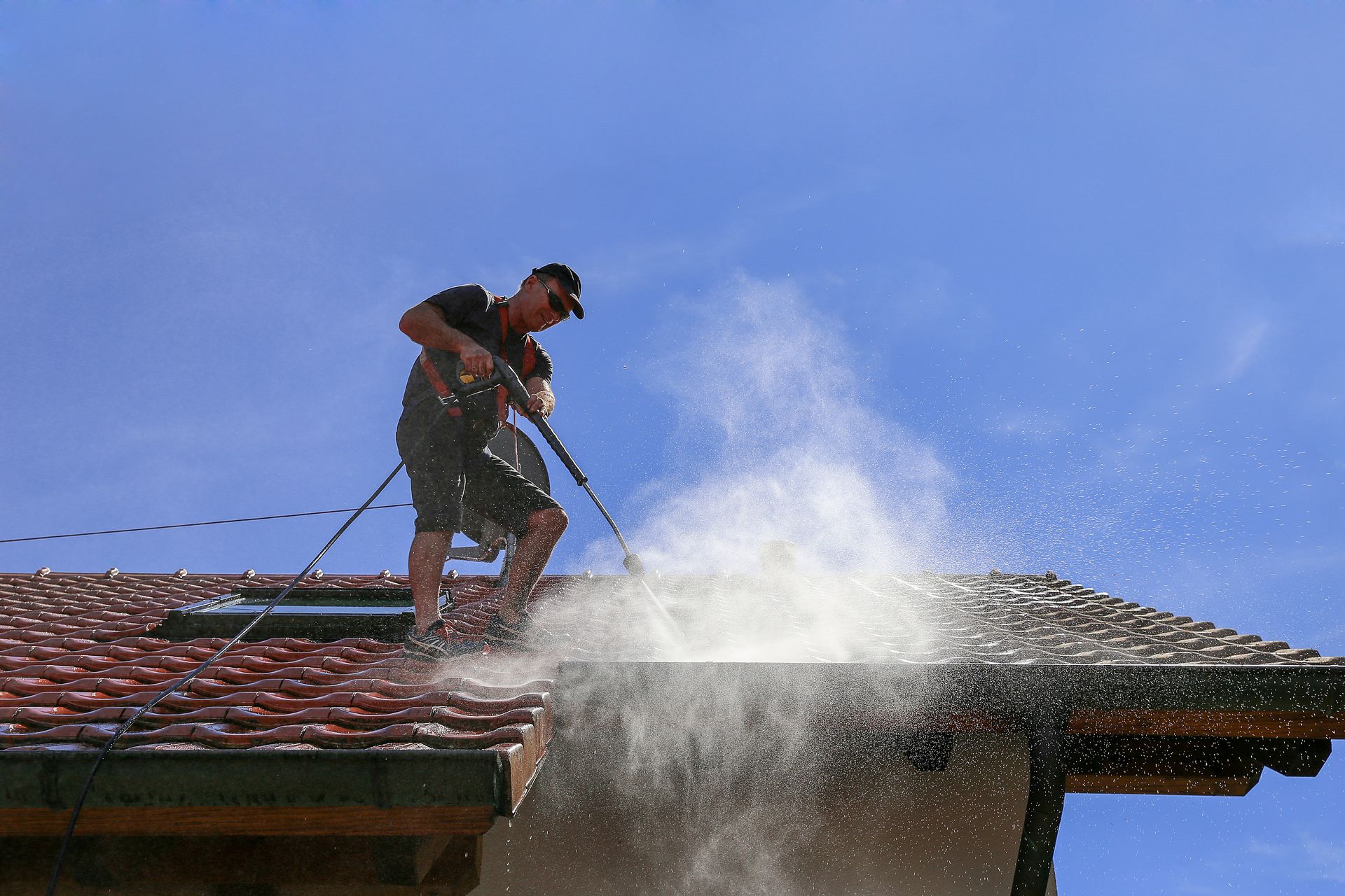 Worker power washing a roof, with white spray against a blue sky.