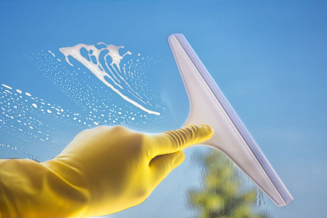 Yellow-gloved hand cleaning a glass window with a squeegee against a blue sky