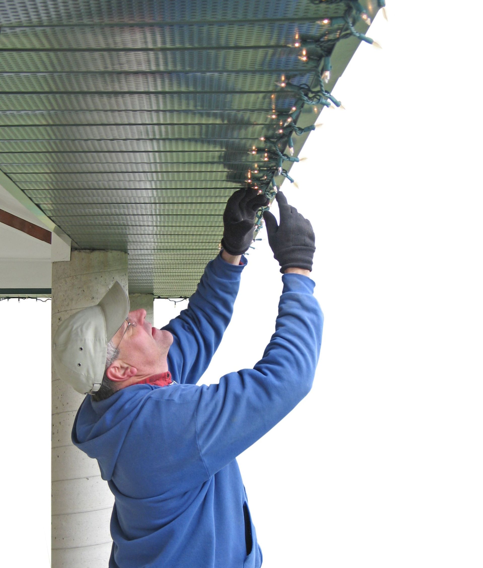 Man in blue jacket and gloves repairing wiring under a corrugated roof overhang.
