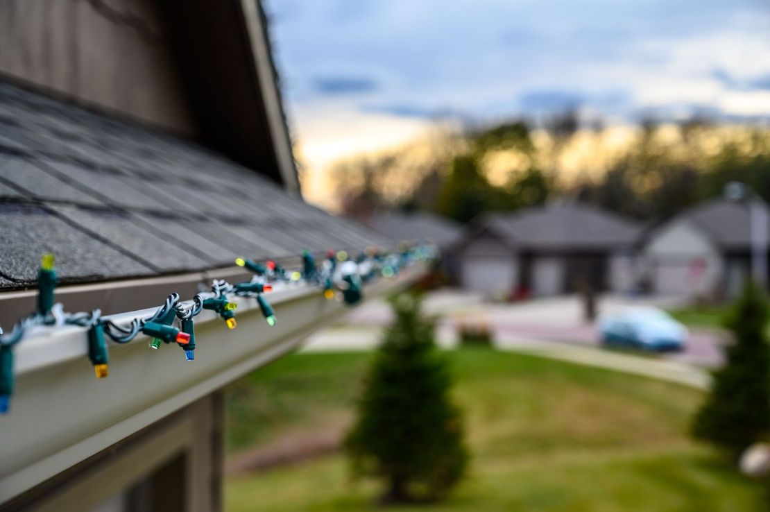 String lights along a roof edge with blurred houses and a sunset sky in the background