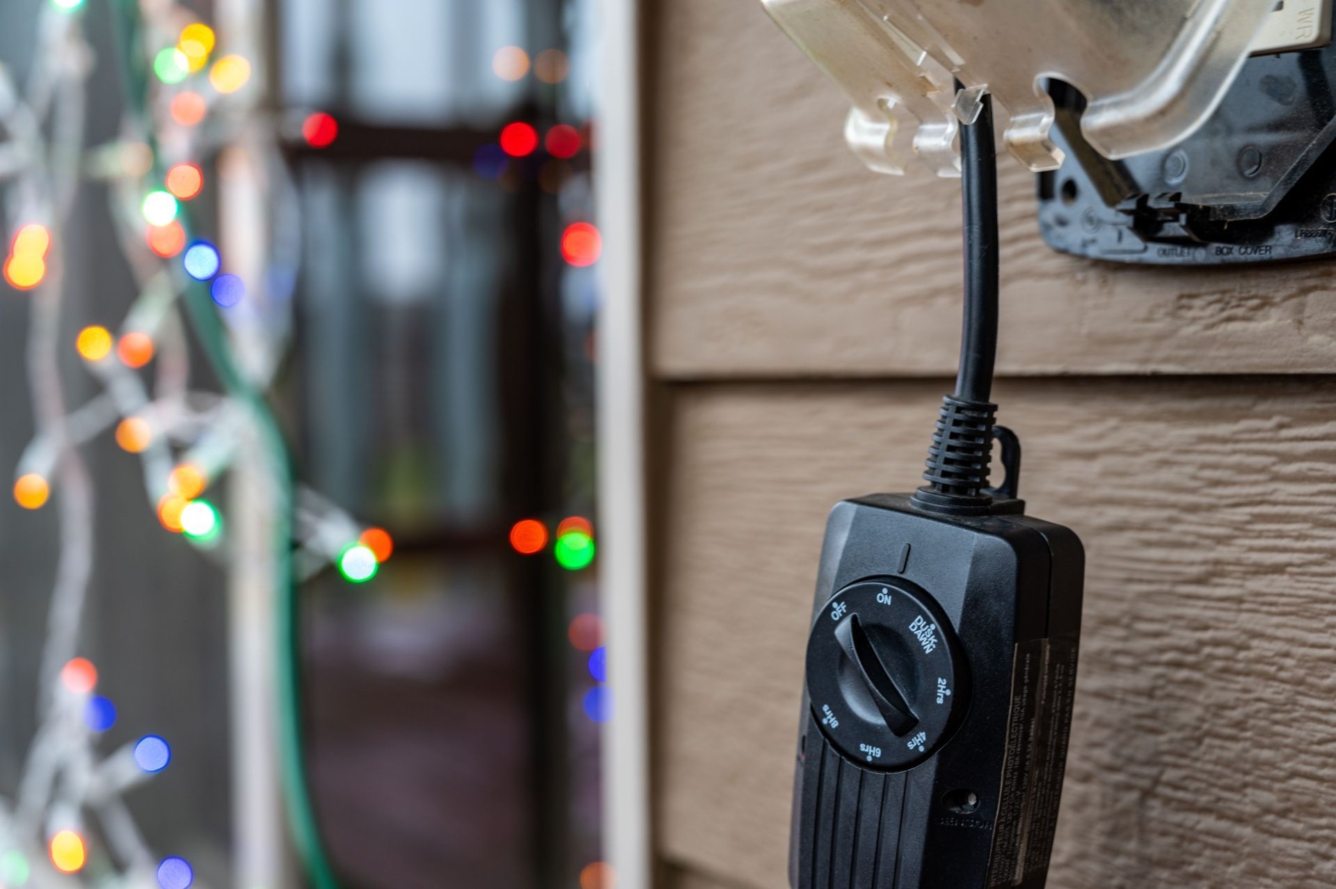 Black power cord plugged into a wall outlet, with blurred colorful string lights in the background.