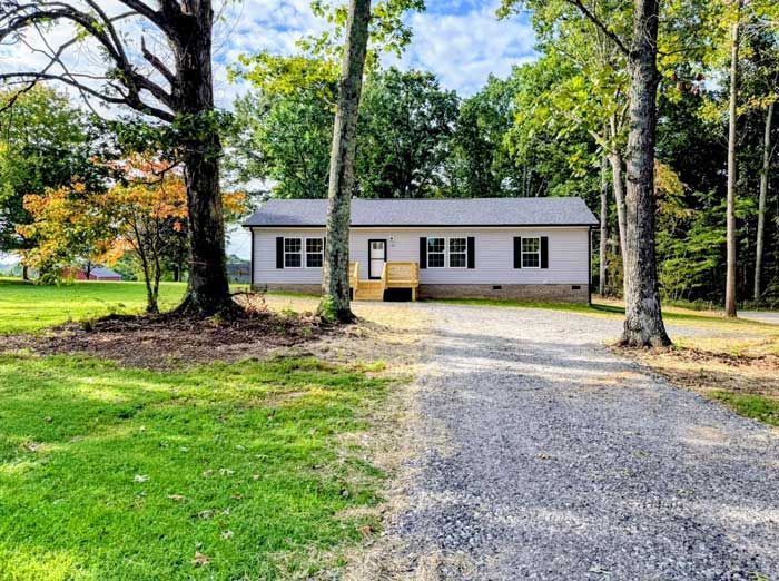 Small white house with a gravel driveway, surrounded by trees and green lawn on a sunny day
