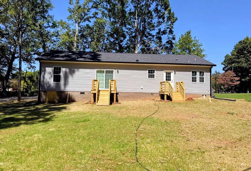 Single-story gray house with two wooden ramps, green lawn, and trees under a clear blue sky