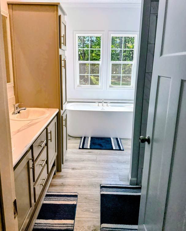 Narrow bathroom with wood vanity, white tub by window, and striped rugs on light wood floor