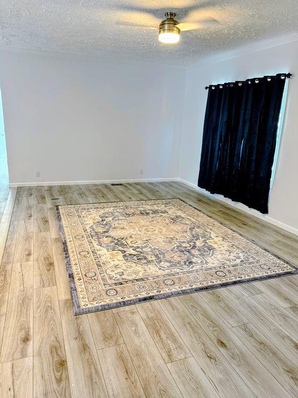 Empty room with light wood floors, a large beige rug, a ceiling light, and black curtains.