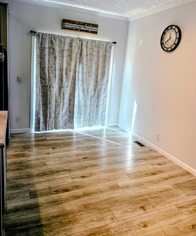 Empty room with wood flooring, curtains over a bright sliding door, and a wall clock on a white wall.