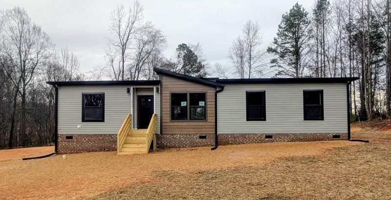 Small beige mobile home with a central porch and stairs, set on a leaf-covered lot with bare trees behind it