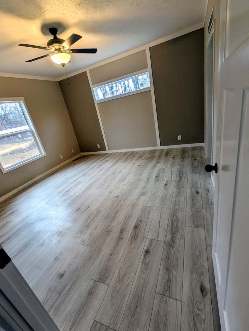 Empty beige bedroom with light wood floors, white ceiling fan, and two windows.