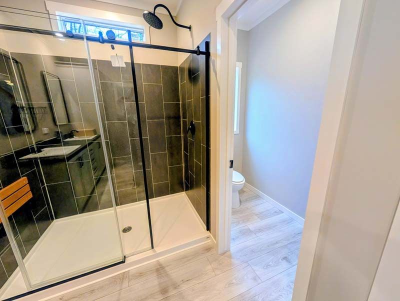 Modern bathroom with glass shower enclosure, vanity sink, and light wood floor leading to an open hallway
