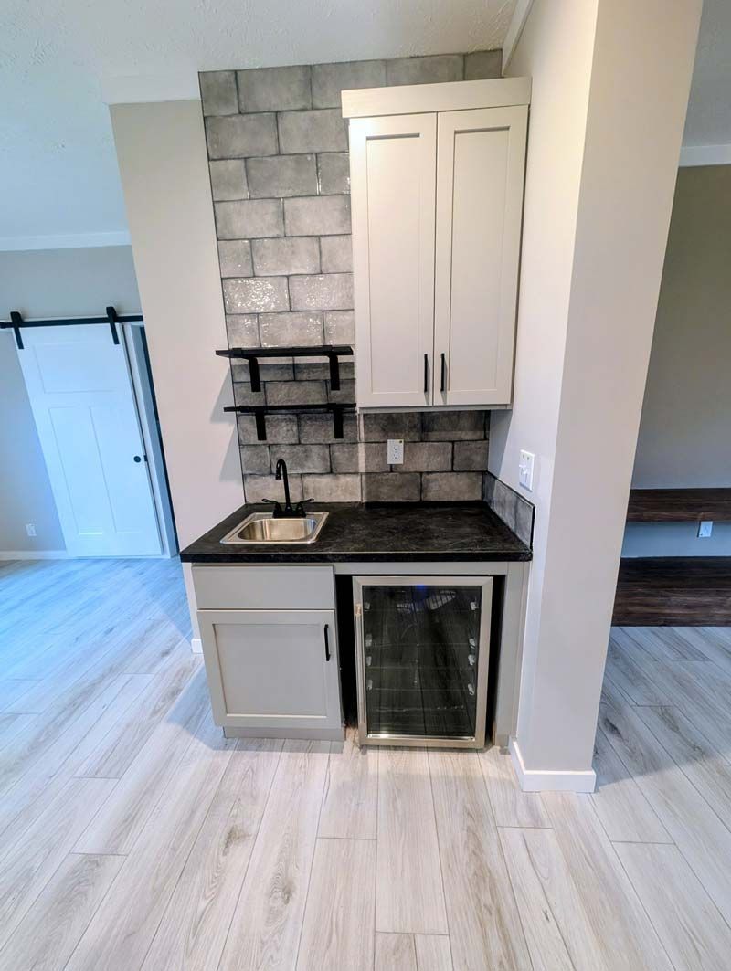 Compact kitchen alcove with sink, black countertop, gray cabinets, and small refrigerator under a white upper cabinet.