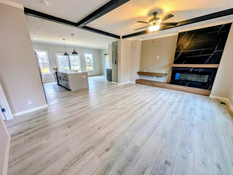 Spacious empty living room with light wood floors, black-trimmed ceiling beams, fireplace, and kitchen view