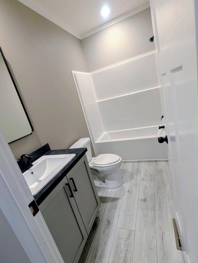 Small modern bathroom with gray vanity, toilet, white shelving, and wood-look tile floor.