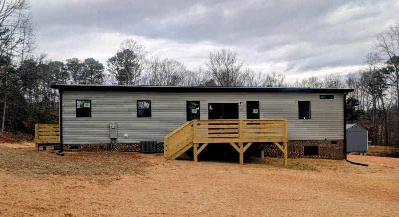 Gray mobile home with a wooden front ramp and steps on a dirt lot under cloudy sky