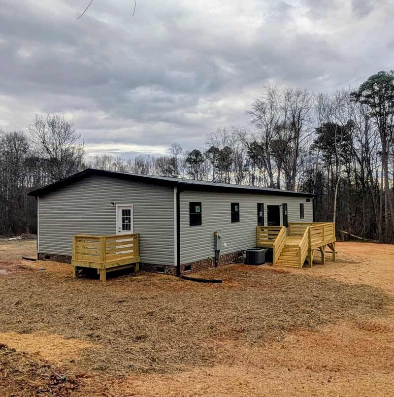 Gray mobile home with wooden steps and deck on a bare lot under cloudy sky