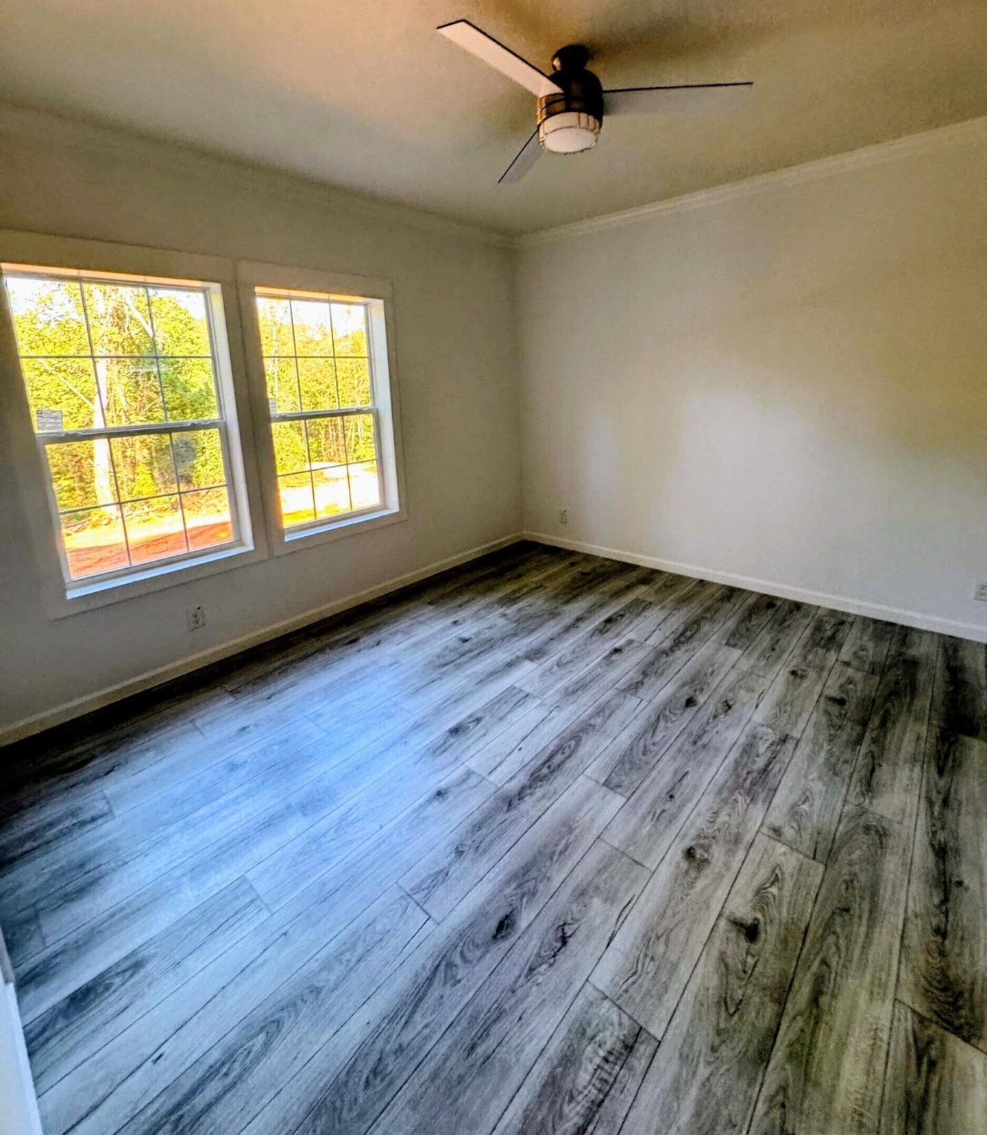 Empty room with gray wood floors, white walls, two windows, and a ceiling fan.