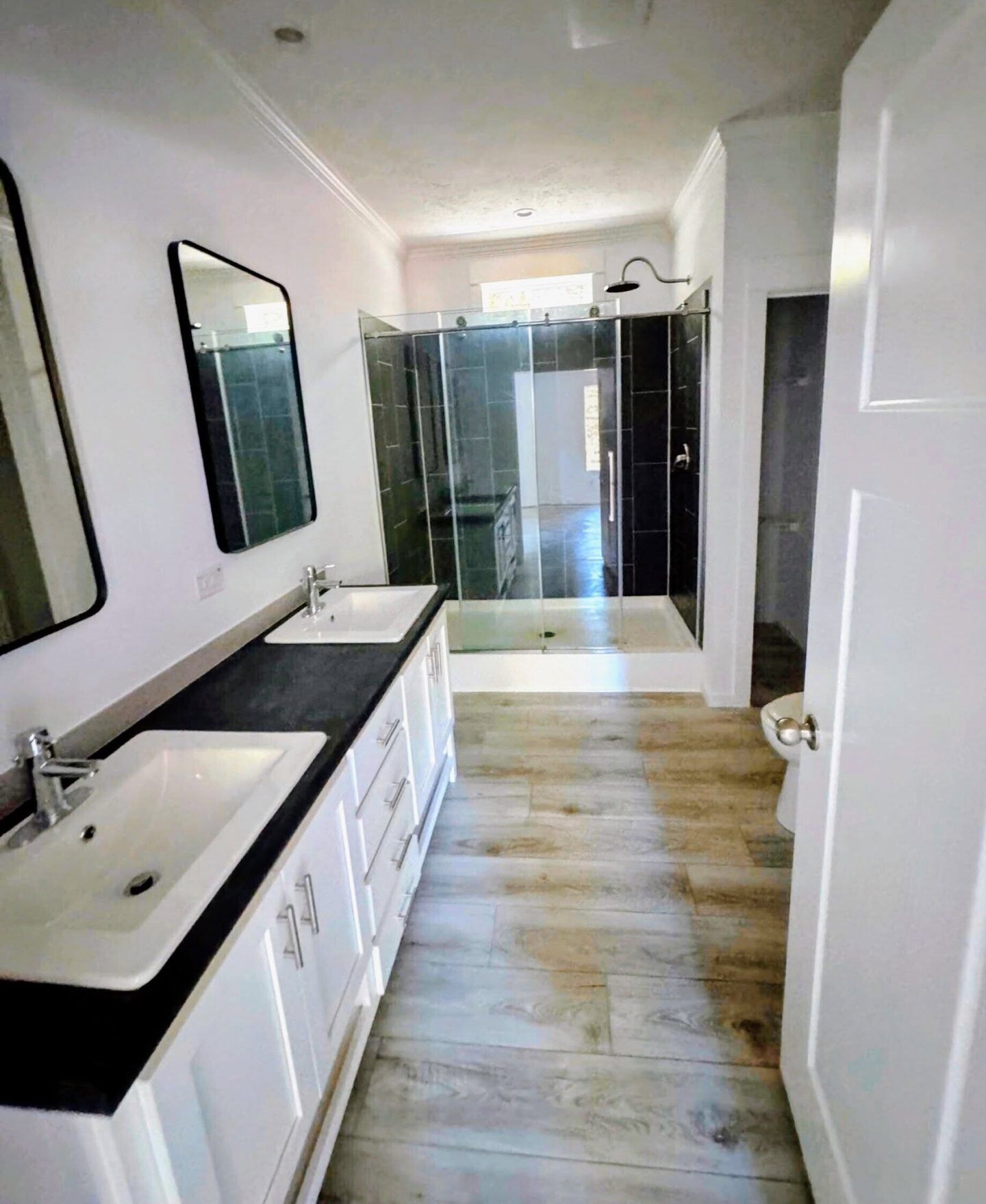 Modern bathroom with double sinks, mirror, black countertop, wood-style floor, and glass-enclosed shower.